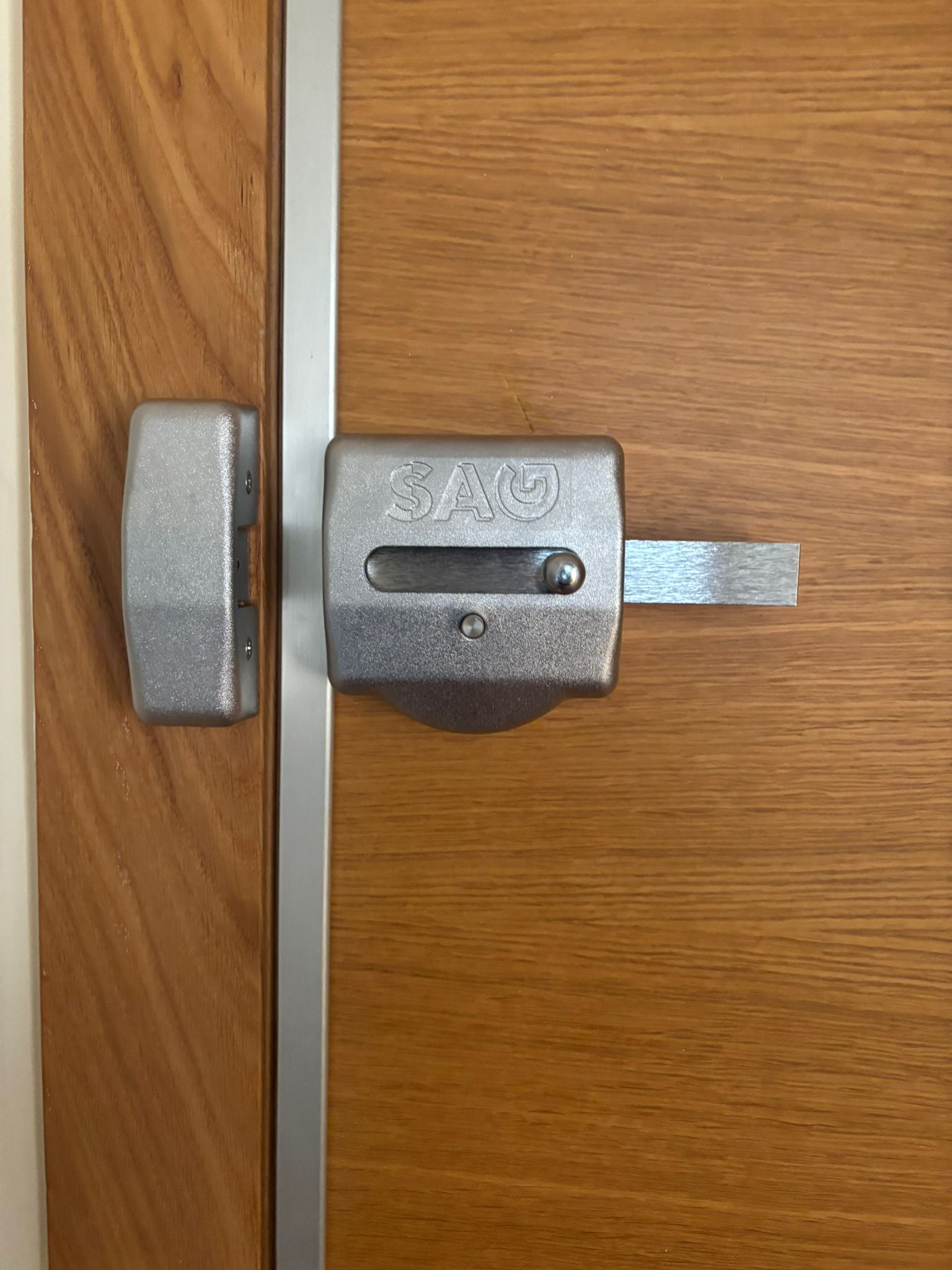 Cerrojo de seguridad de sobreponer SAG en acero gris con pestillo extendido instalado en puerta de madera clara