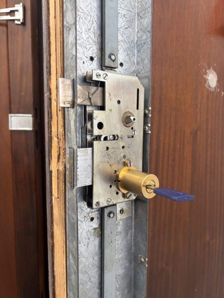 Mecanismo interior de cerradura multipunto al descubierto con bombín nuevo en latón y llave azul durante sustitución en puerta blindada